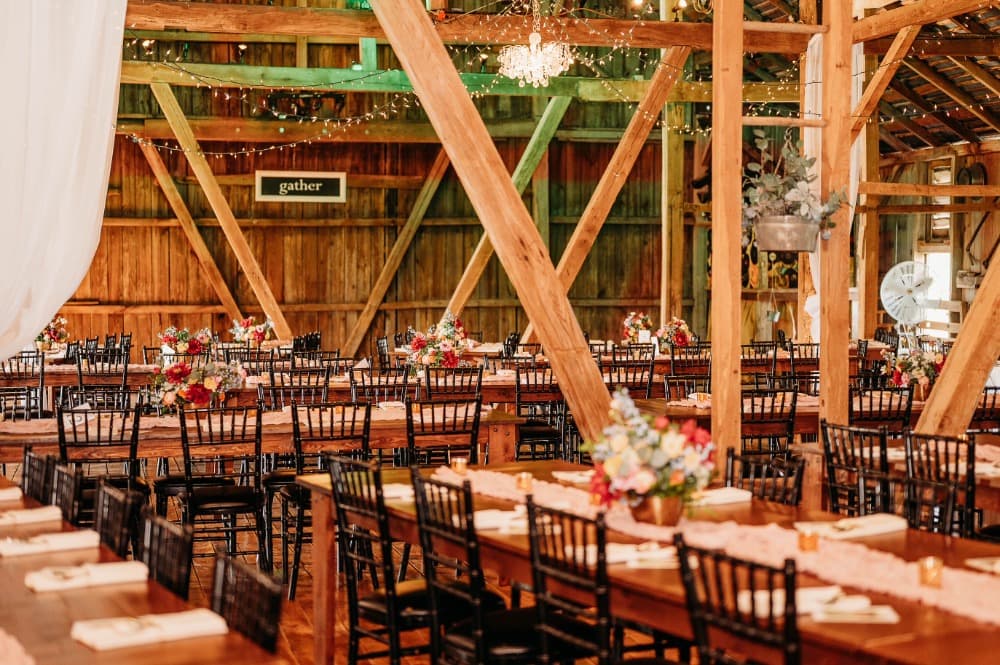 Long wooden farm tables and black chiavari chairs set for dinner beneath the timber beams and string lights of the main barn.
